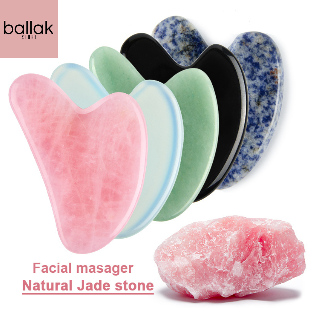 A collection of natural stone Gua Sha massagers in various colors including black, blue, green, pink, and purple, alongside a raw piece of pink stone. The massagers are designed in a heart shape with a smooth, curved surface.