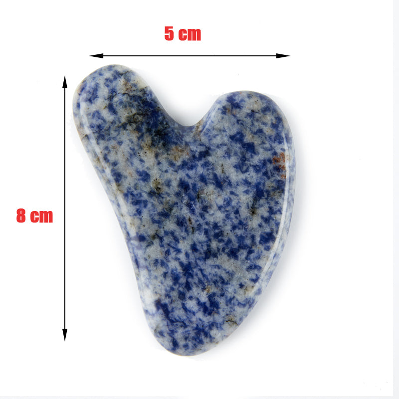 A collection of natural stone Gua Sha massagers in various colors including black, blue, green, pink, and purple, alongside a raw piece of pink stone. The massagers are designed in a heart shape with a smooth, curved surface.