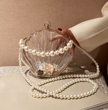 Shell-shaped handbag with pearl strap held by a hand against a neutral background